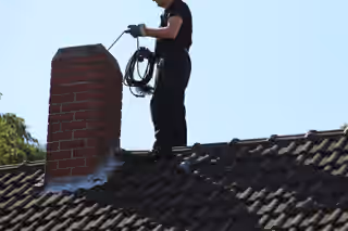 Ramoneur debout sur un toit, hérisson et corde de ramonage à la main, au-dessus d’une souche en brique